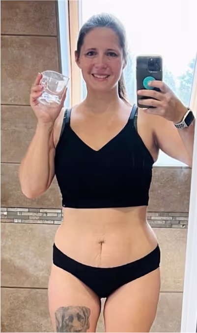 Eine lächelnde Frau in Sport-BH und Unterwäsche macht ein Selfie im Spiegel und hält ein Glas.
