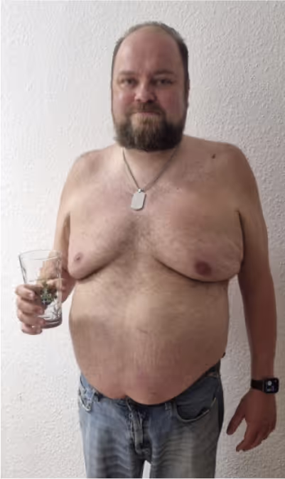  Ein lächelnder Mann ohne Hemd, in Jeans und mit Bart hält ein Glas in der Hand.
