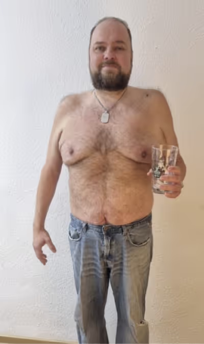  Ein lächelnder Mann ohne Hemd, in Jeans und mit Bart hält ein Glas in der Hand.