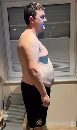 Ein Mann ohne Hemd und in Shorts steht seitlich vor einem Fenster.