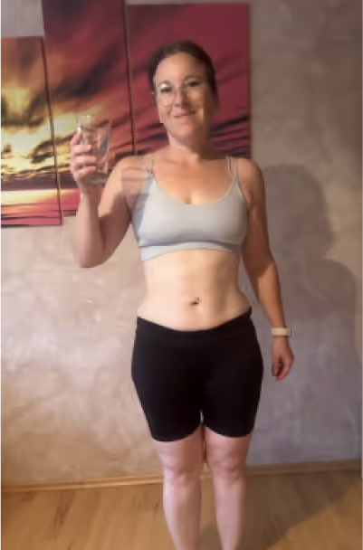 Eine lächelnde Frau in Sportkleidung hält ein Glas Wasser in der Hand.