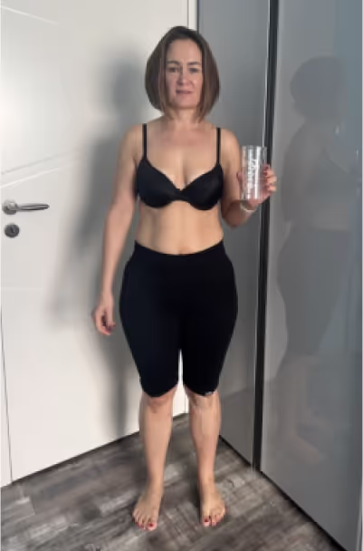 Eine Frau in schwarzem Sport-BH und Shorts steht vor einer weißen Tür und hält ein Glas.