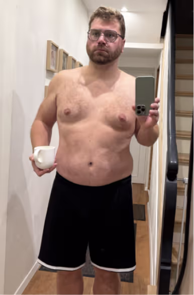Ein Mann ohne Hemd, in Shorts und mit Brille macht ein Selfie im Spiegel und hält eine Tasse.